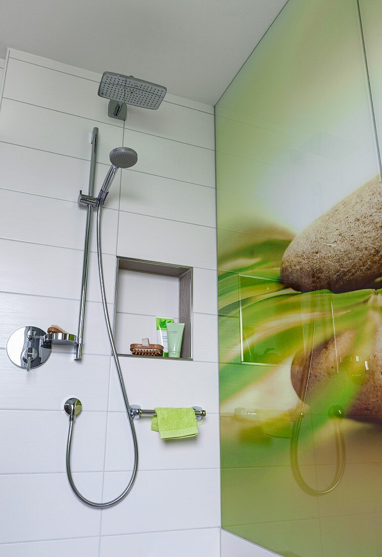 A hand-held shower head and a rain shower head on a white tiled wall with a partially visible printed glass back splash with a natural design in a shower
