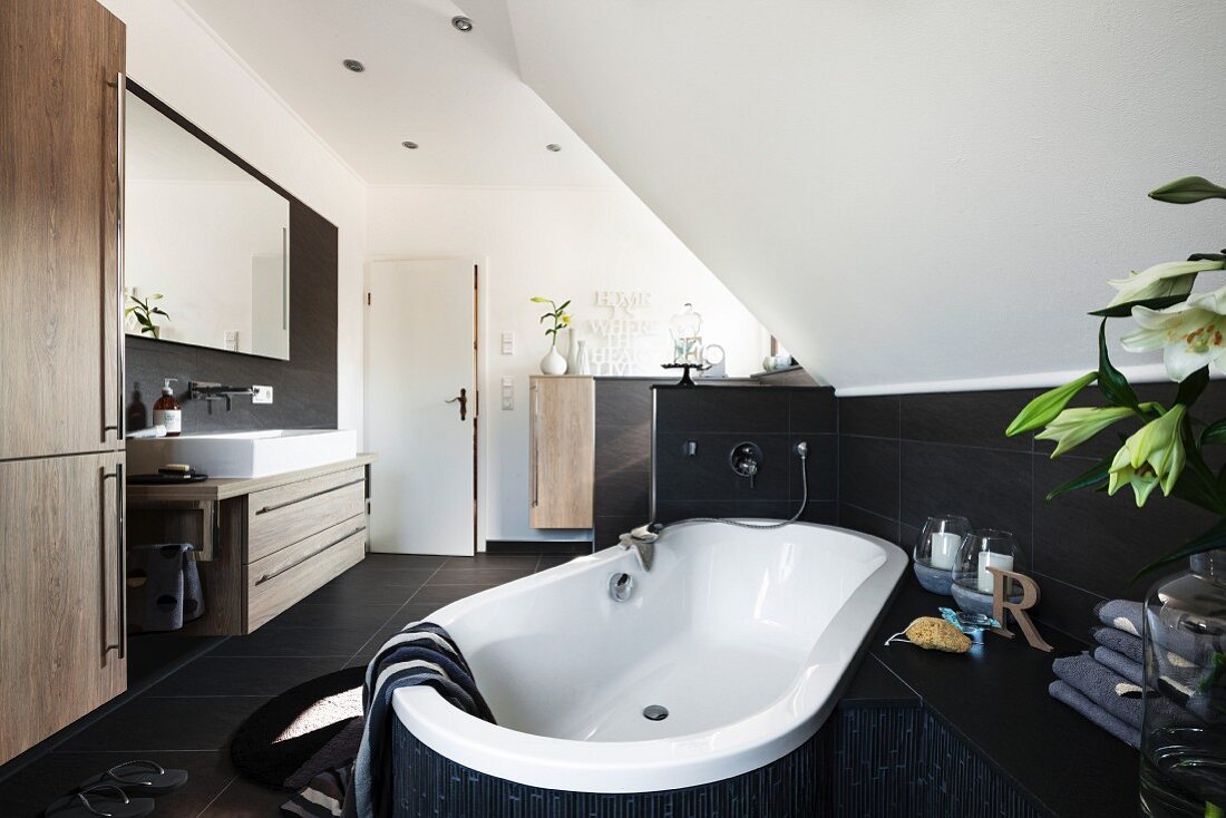 A bathtub positioned at an angle in the room under a vaulted roof with brown tiles halfway up the wall opposite a trough-style washstand and a large mirror on the wall in a modern bathroom