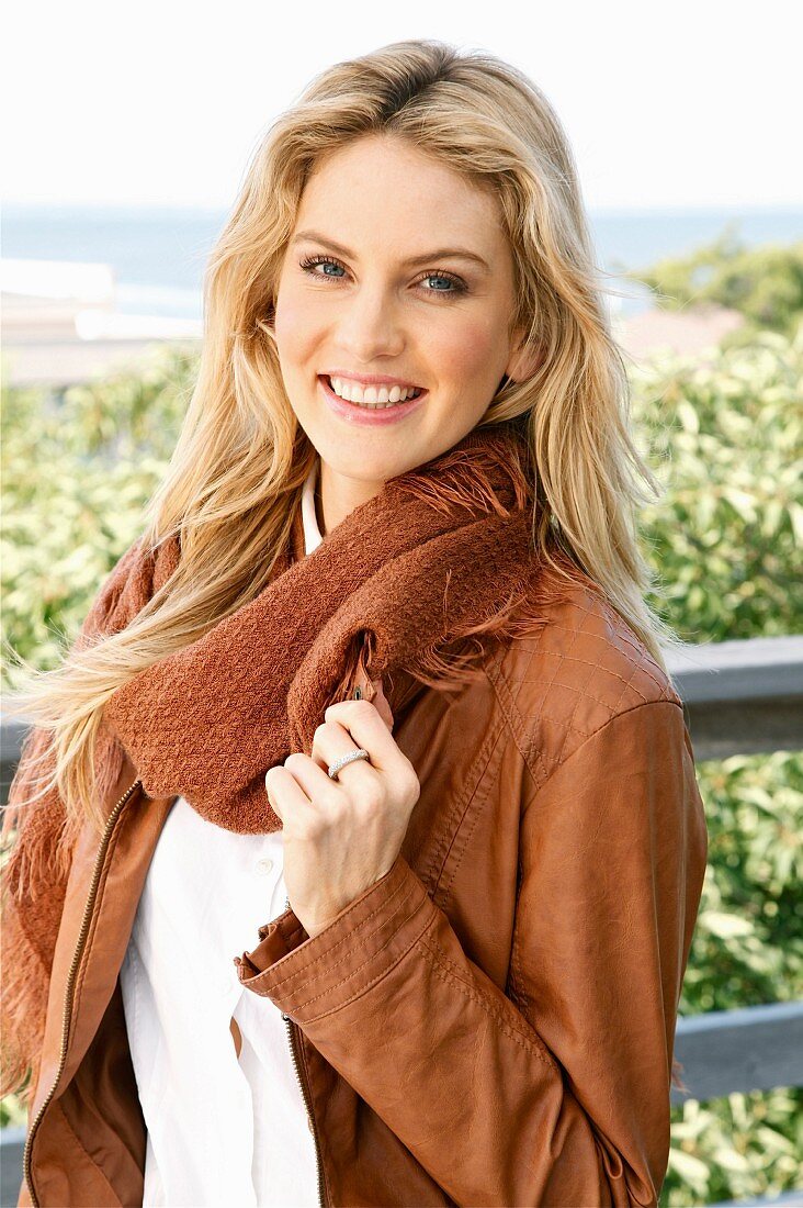 A young blonde woman outside wearing a brown leather jacket, a white shirt and a scarf