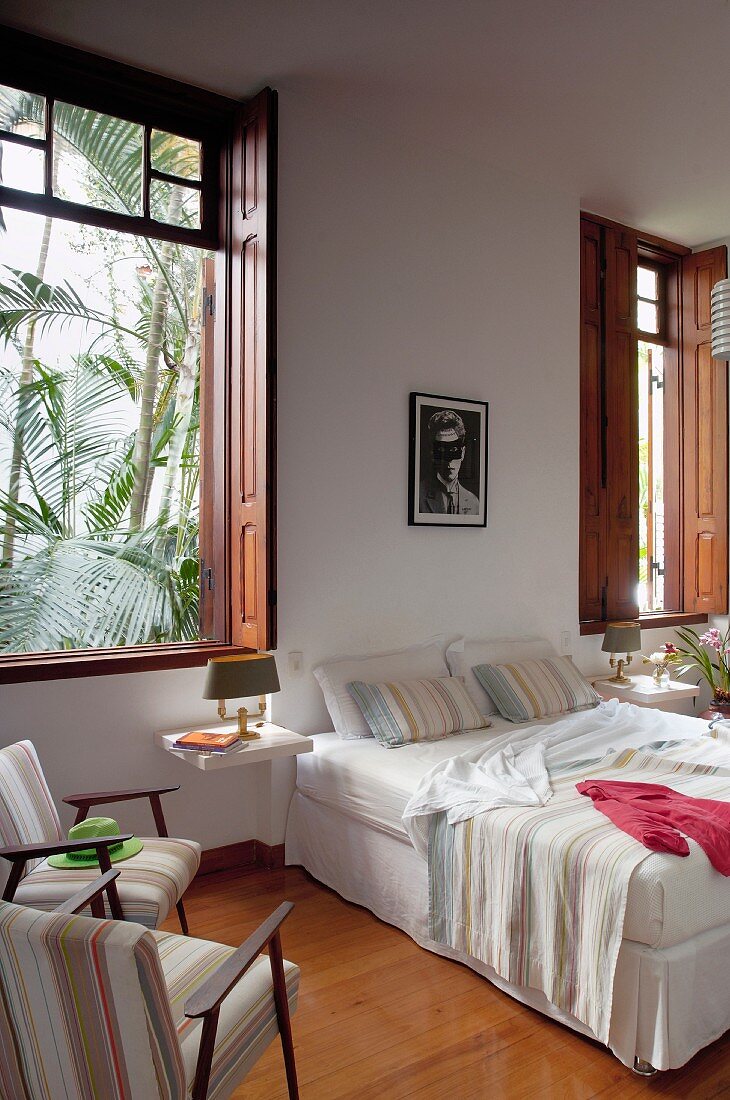 Comfortable double bed against wall, armchairs next to window with wooden interior shutters and view of palm trees