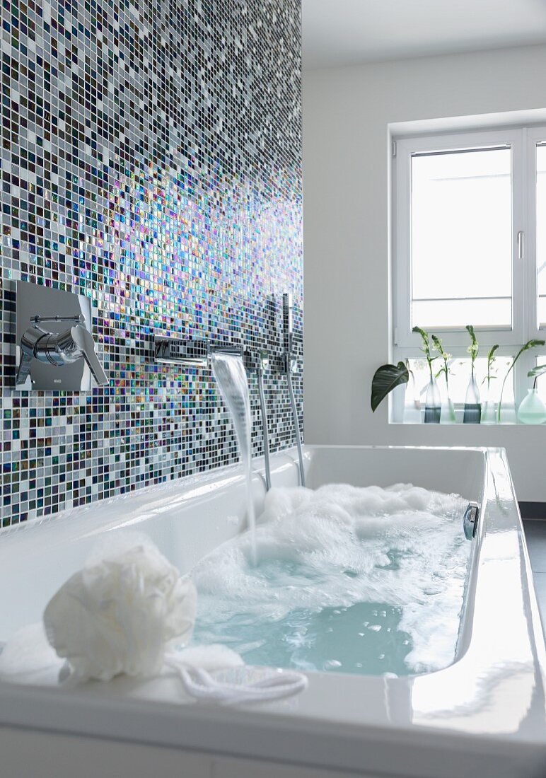 A shimmering mosaic tiled wall behind a bathtub with a bubble bath running