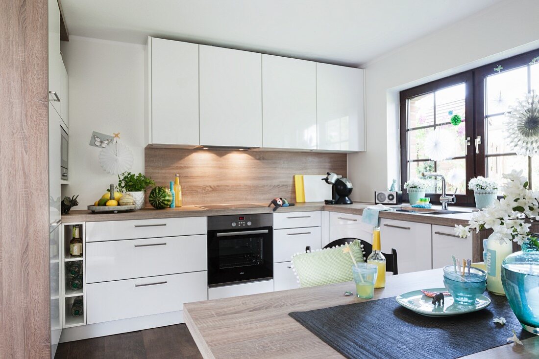 A white modern fitted kitchen with shiny white fronts and a wooden look dining table in the foreground