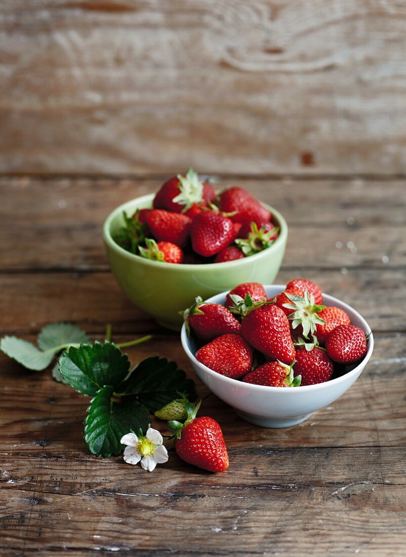 Bowls of fresh strawberries with leaves and flowers on a wooden surface