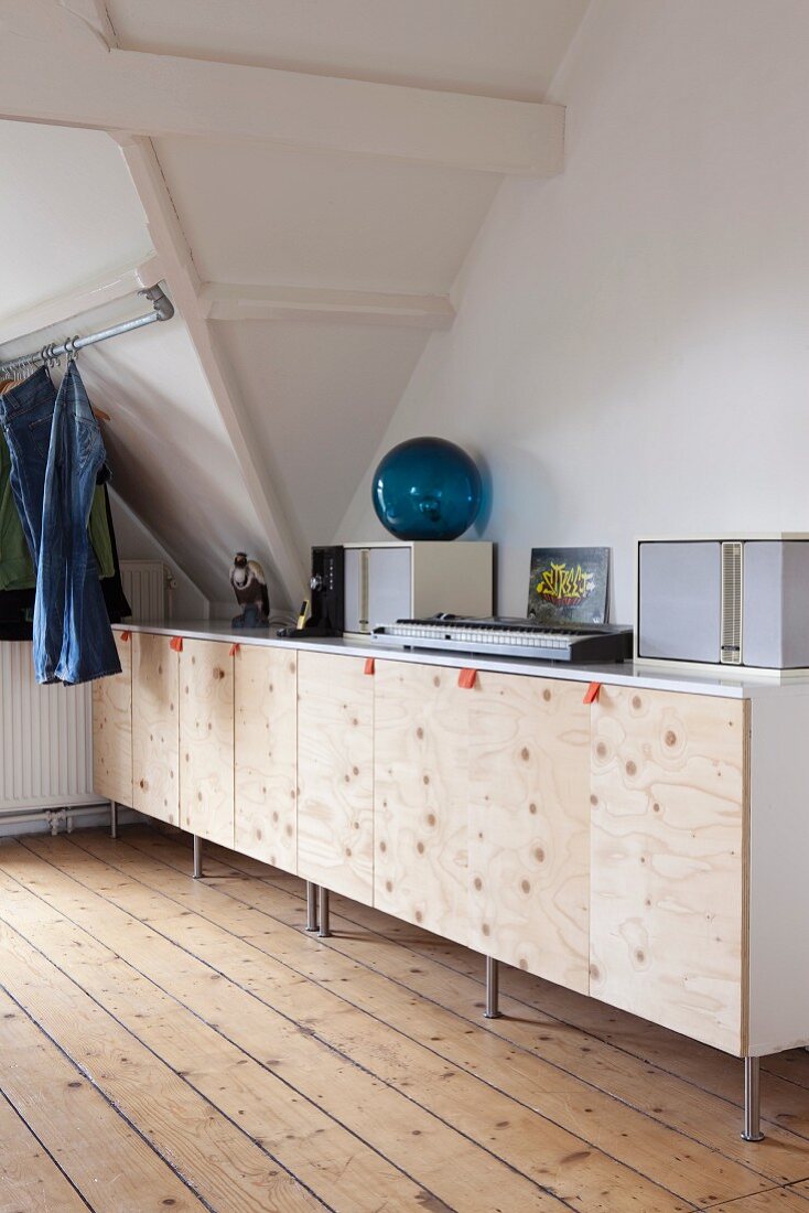 Keyboard and speakers on sideboard with plain wooden doors and fabric loop handles in attic room with jeans hanging from metal rod
