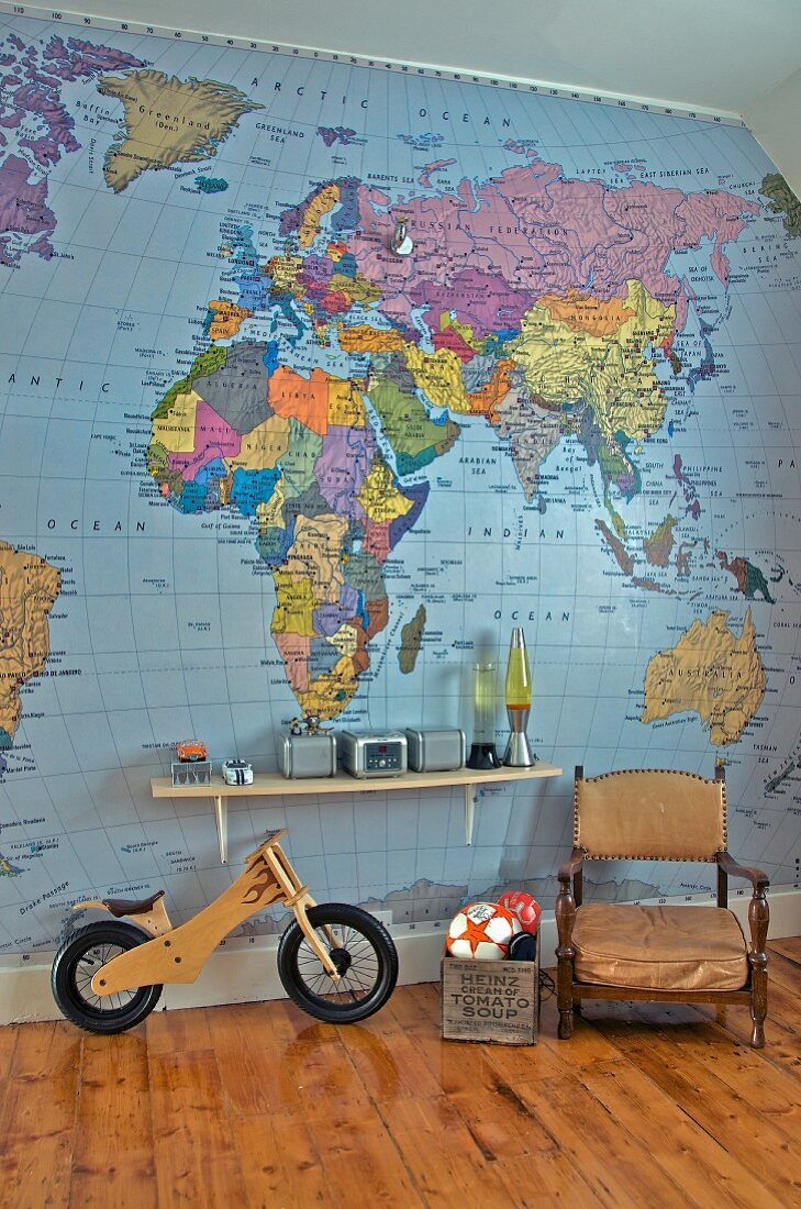 Antique armchair next to toy box and balance bike below bracket shelf on wall papered with map of world