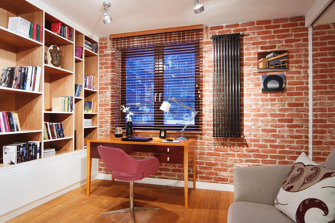 Simple workspace below window; pink swivel chair and wooden desk against brick wall, wooden, fitted shelving with painted fronts to one side