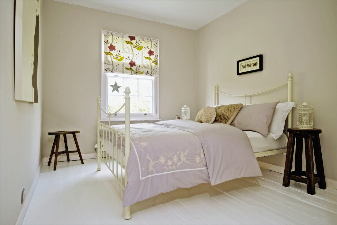 Double bed with white, antique metal frame in bedroom painted pale grey with patterned roller blind on lattice window