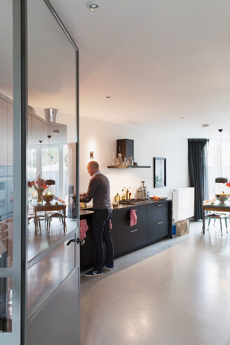 View through open door into kitchen with black base units, concrete floor and man stood at counter