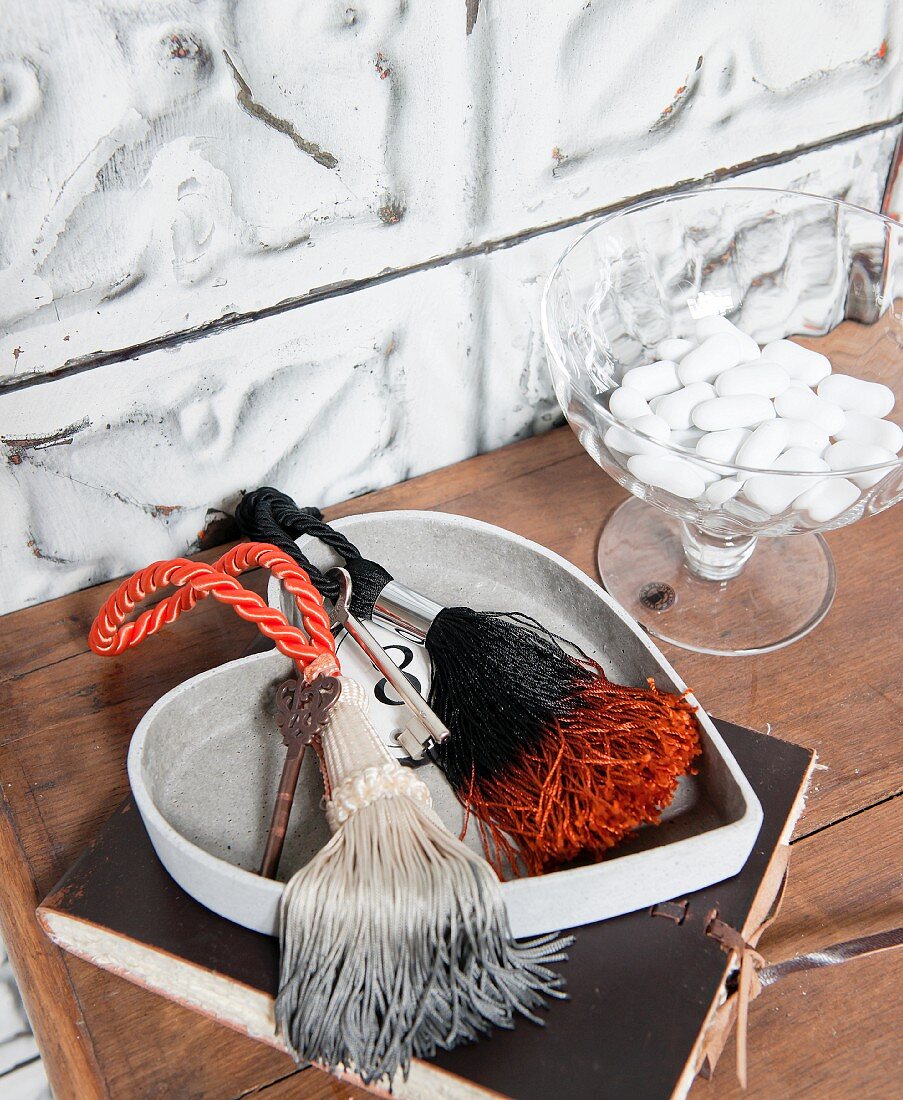 Colourful dip-dyed cords and tassels in heart-shaped china dish and glass dish of sweets on wooden cabinet