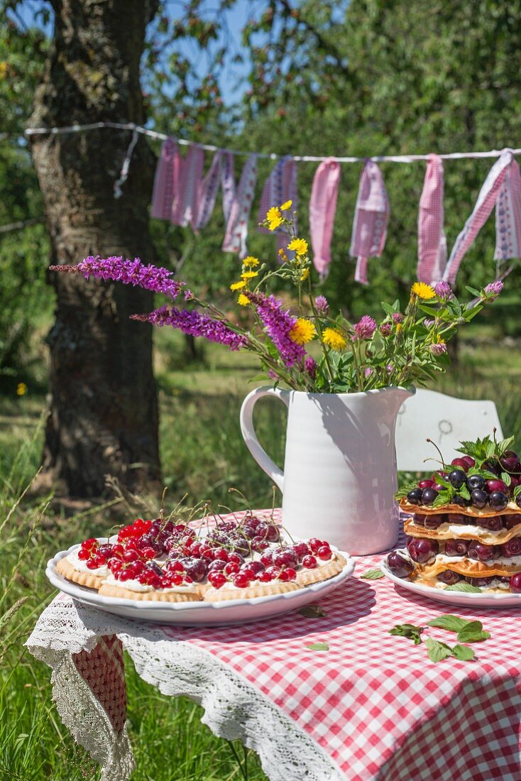 Tartlets and waffles with fresh cherries and a bunch of wild flowers in a jug on a garden table with a checked table cloth
