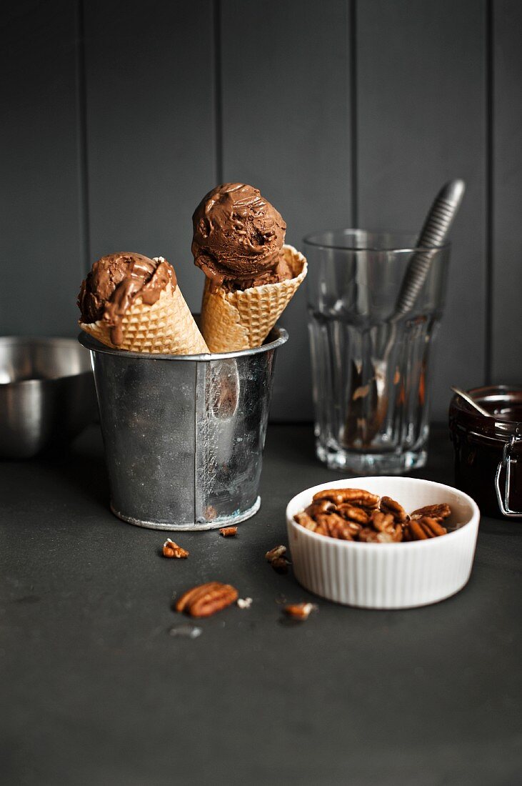 Two cones of chocolate ice cream in a metal bucket next to a bowl of pecan nuts