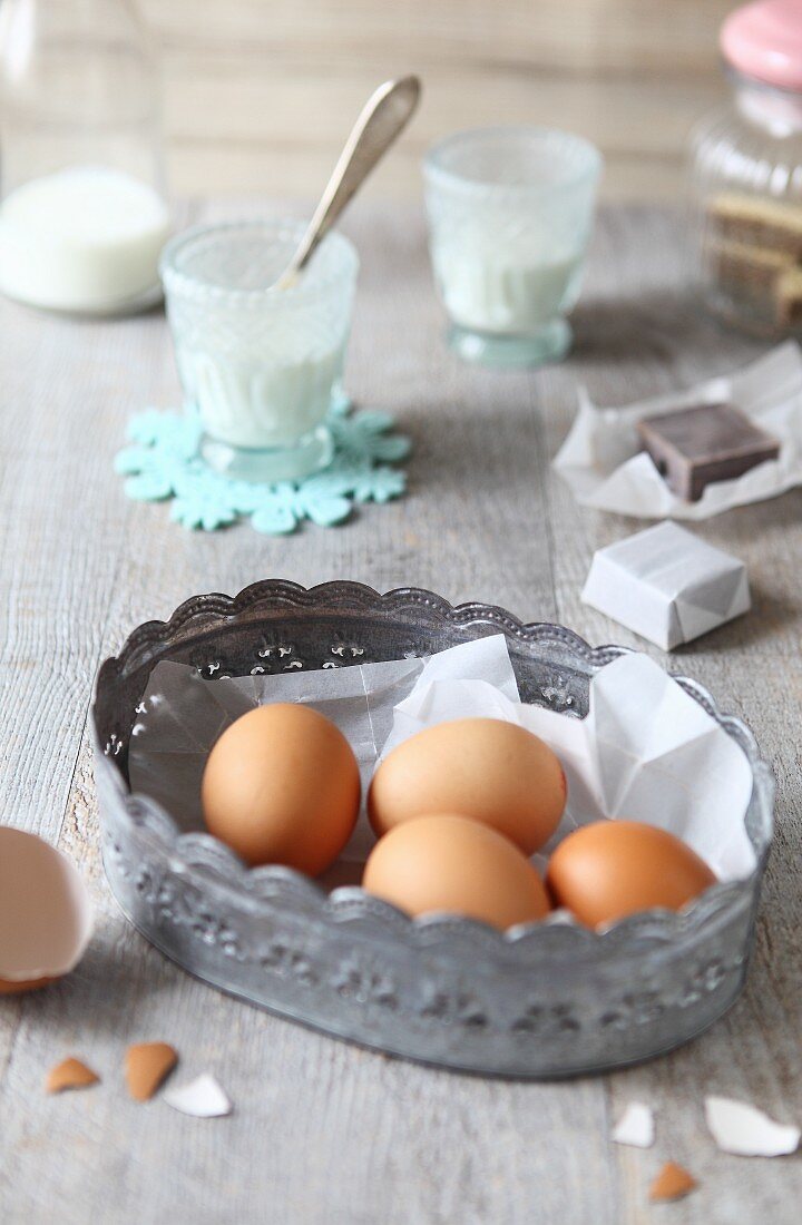 Vier braune Eier im Metallkörbchen auf Holzplatte sowie verschiedene Backzutaten