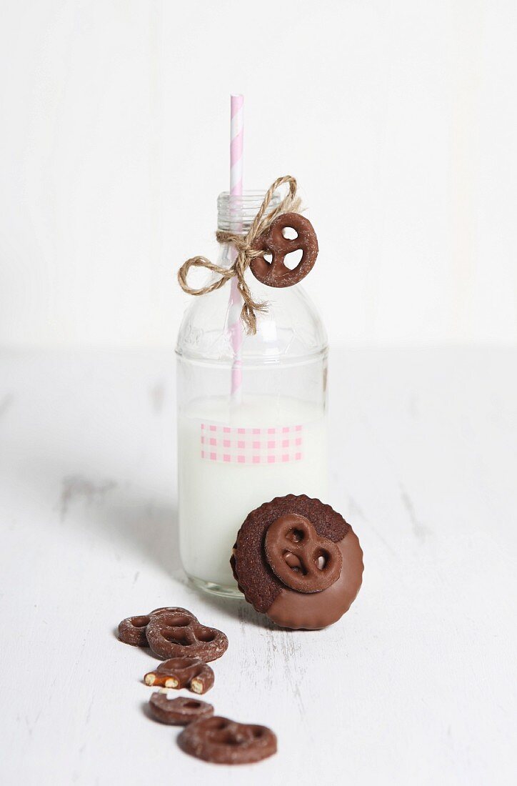 A bottle of milk with a straw and chocolate pretzels on a white wooden surface