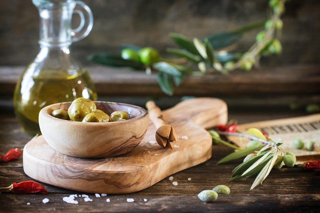 Grüne Oliven in Olivenholzschale und eine Flasche Olivenöl auf altem Holztisch