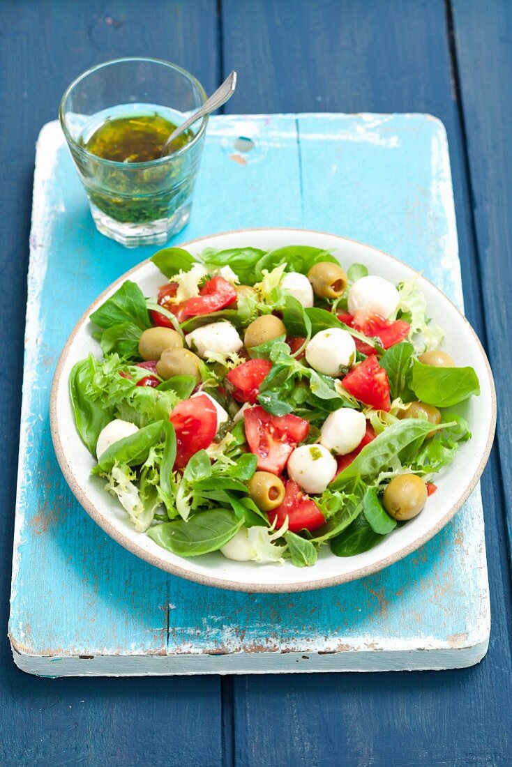 Gemischter Salat mit Mozzarella und grünen Oliven; dahinter eine Kräutervinaigrette im Glas
