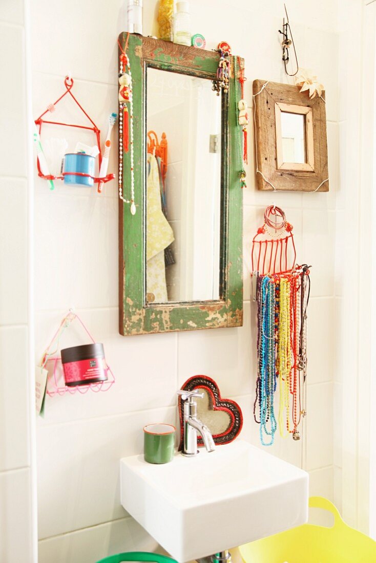 Jewellery hung from mirror frame with peeling paint and wire basket of bead necklaces and toiletries above small sink