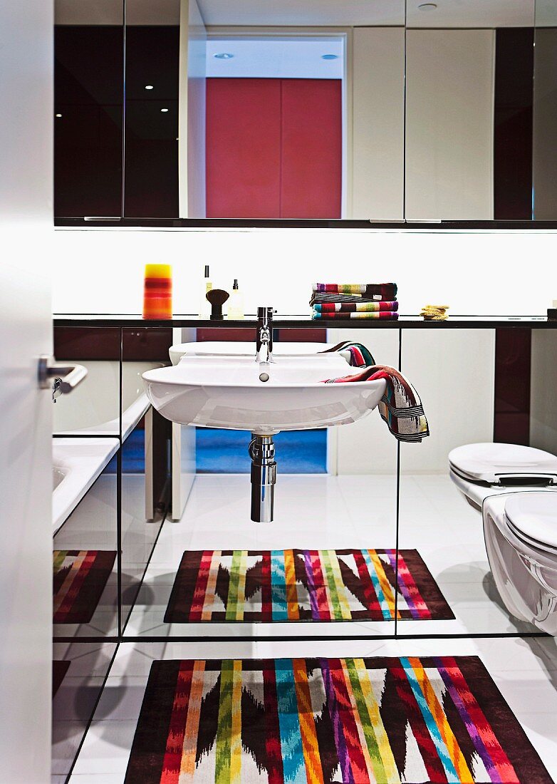 Brightly coloured rug reflected in mirrored wall behind sink and wall-mounted toilet below mirrored cabinet