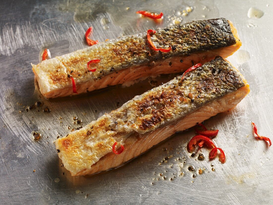 Two crispy grilled salmon fillets with black pepper and chilli strips on a metal surface