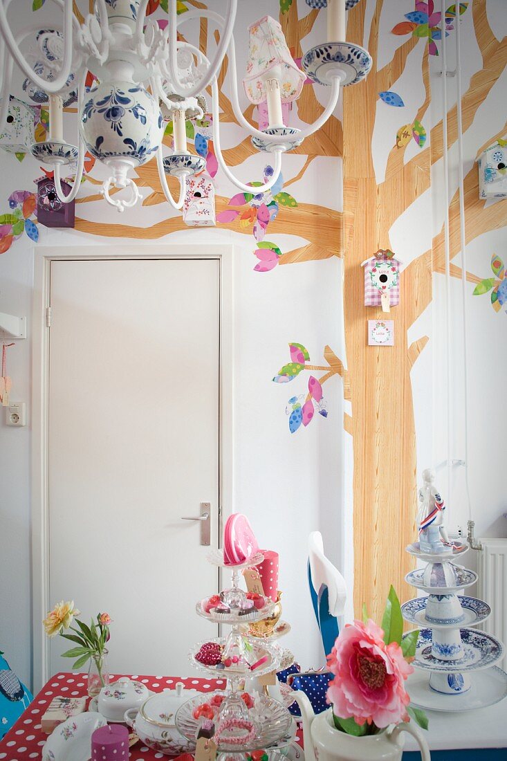 Multi-armed chandelier above table decorated with romantic charm; nesting boxes hung on wall with tree mural