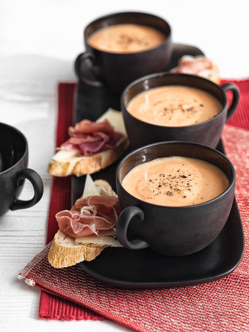 Cold tomato soup in black cups with rolls topped with Serrano ham and Manchego cheese