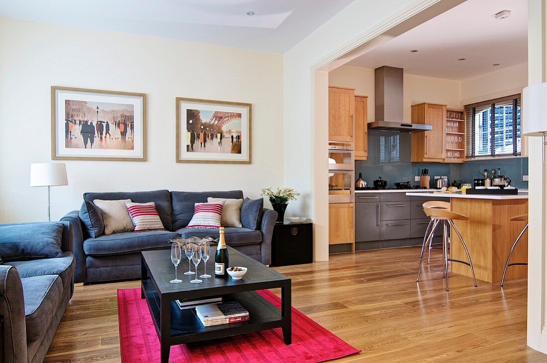 Champagne flutes on table on raspberry rug, grey sofas, open-plan kitchen with pale wooden furniture and grey base cabinets