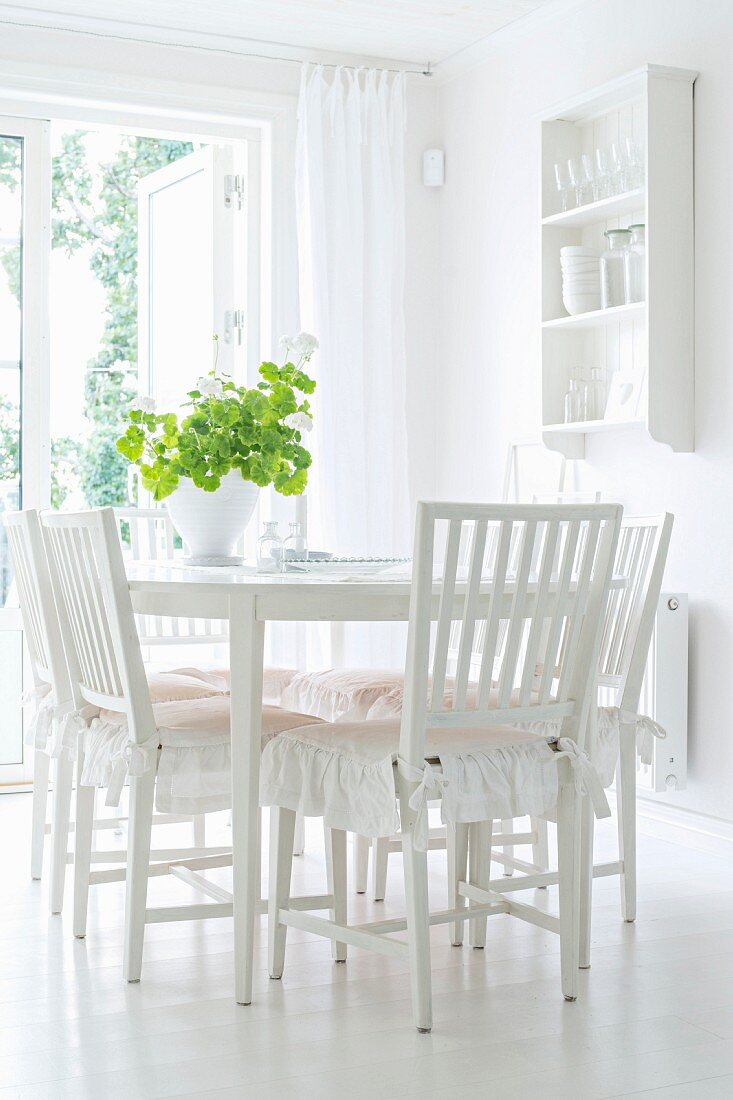 White dining room with frilled cushions on wooden chairs and crockery shelves on wall
