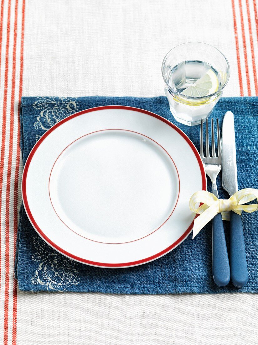 An empty white plate with a red rim on a blue placemat in the glass of water