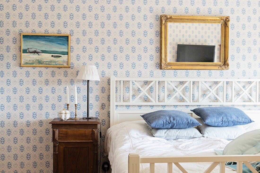 Doppelbett mit Kopfteil aus weißem Holz in ländlichem Schlafzimmer, Bild und Goldrahmenspiegel an weiss-blau gemusterter Tapete