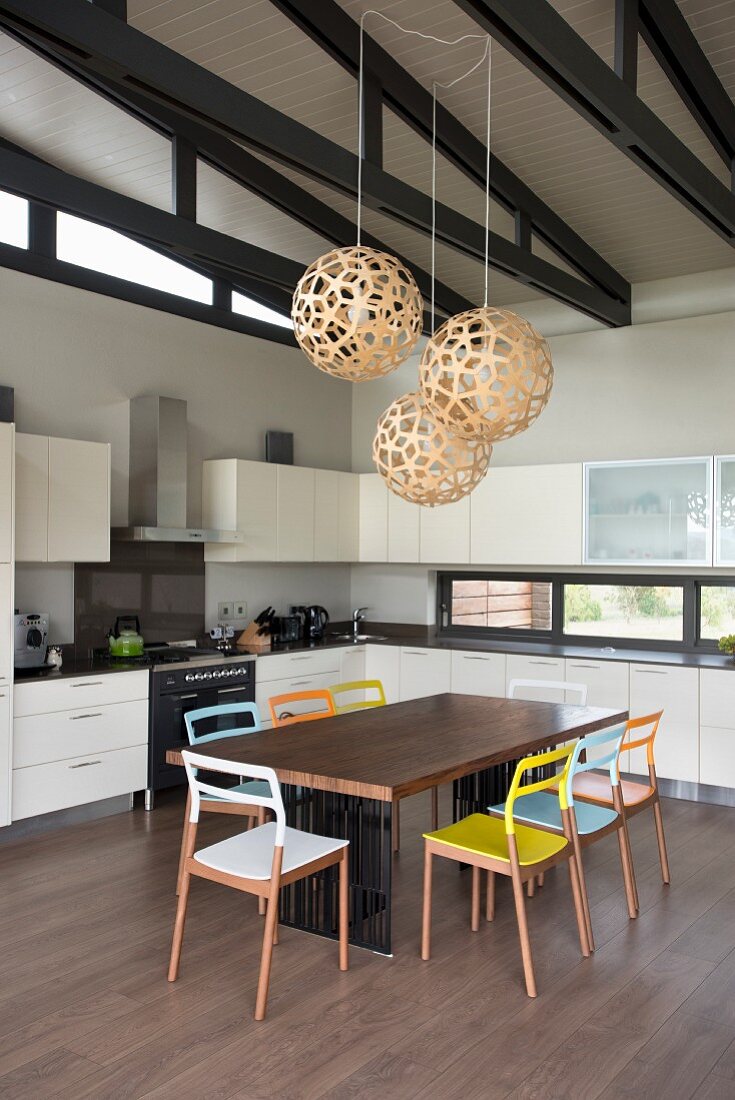 Wooden chairs with colourful backrests and seats around table in white fitted kitchen with spherical pendant lamps