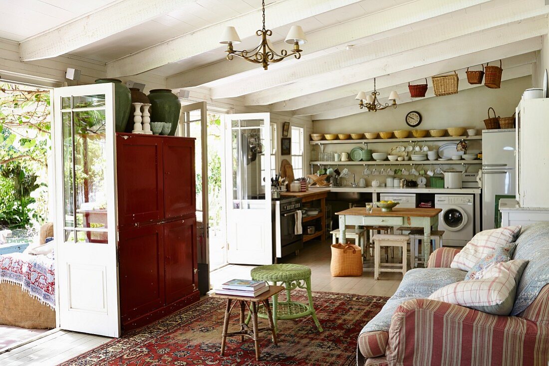 Rustic interior with white, wood-beamed ceiling, couch with scatter cushions opposite open terrace doors and kitchen in background