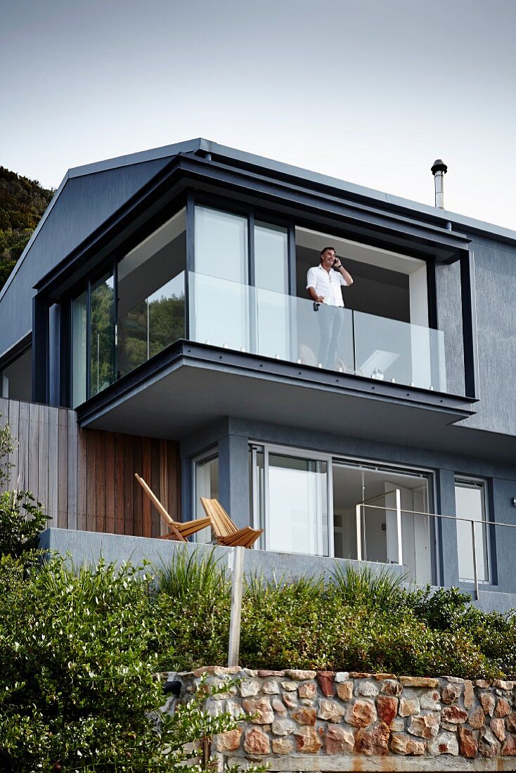 Contemporary house with grey-painted facade; man standing behind glass balustrade of open terrace windows of upper storey