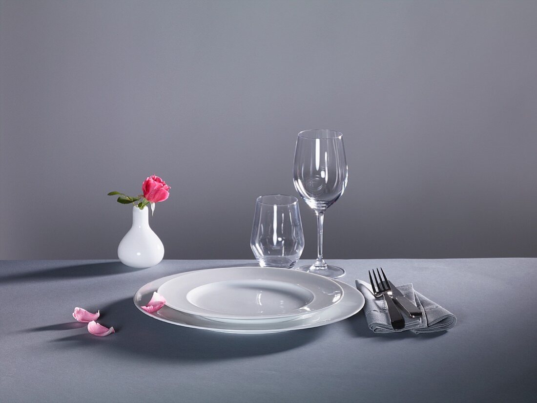 A place setting against a grey background: plates, cutlery, empty glasses and a flower vase