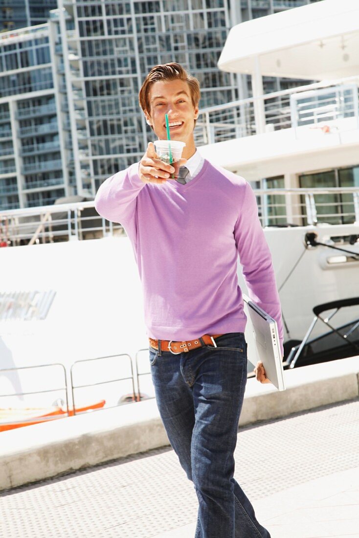 A young businessman holding a drink and a laptop walking down a road with an office block in the background