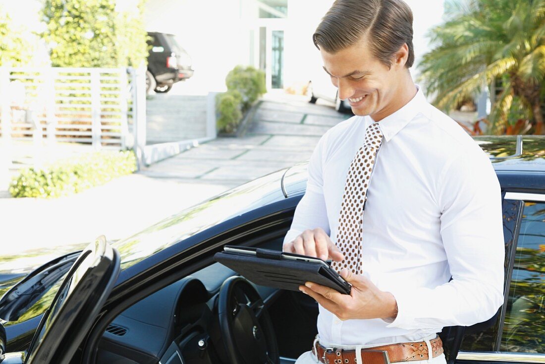 A young businessman standing next to a car holding a tablet computer