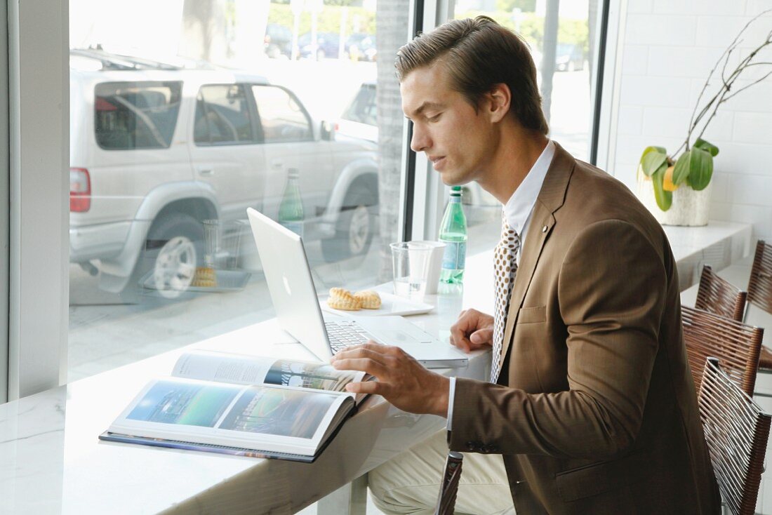 A young businessmen wearing a brown blazer sitting in an office in front of a laptop and an open book