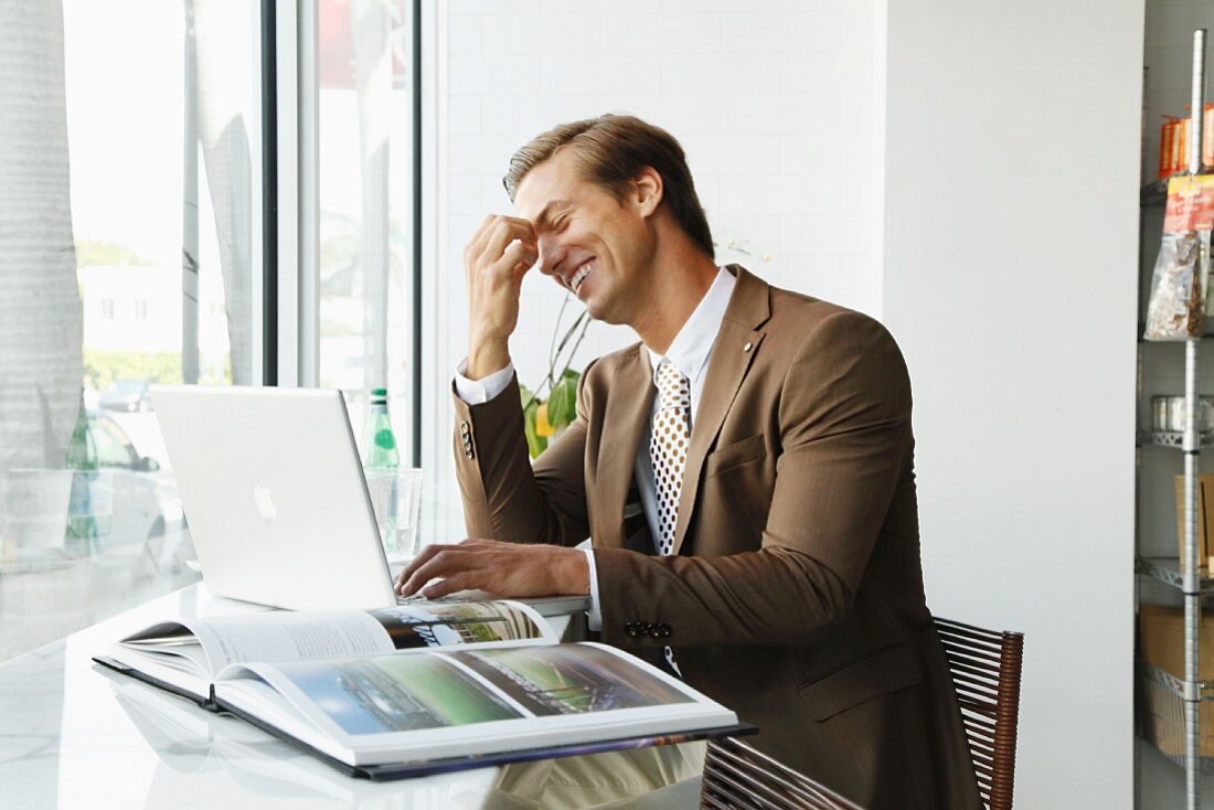 A young businessman wearing a brown blazer sitting in an office in front of a laptop and an open book