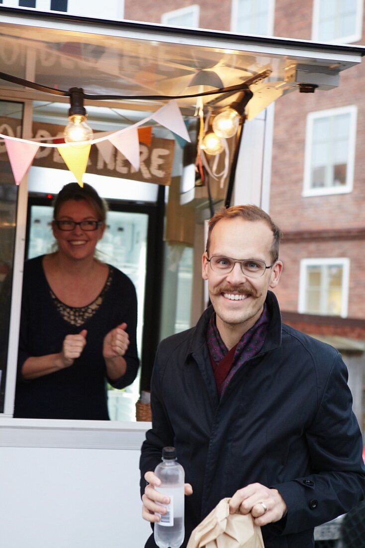 A happy man in front of a mobile food unit holding a bottle of water and a sandwich in a paper bag with a smiling sales assistant in the background