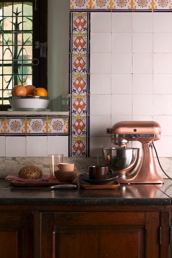 Old, country-house kitchen with vintage-style mixer on rustic base cabinet and decorative borders of colourful patterned tiles on wall