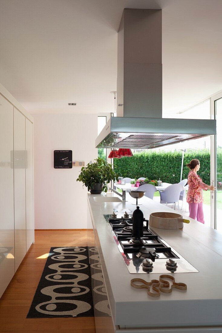 Kitchen counter with integrated gas hob below stainless steel extractor hood; woman standing in terrace windows in background