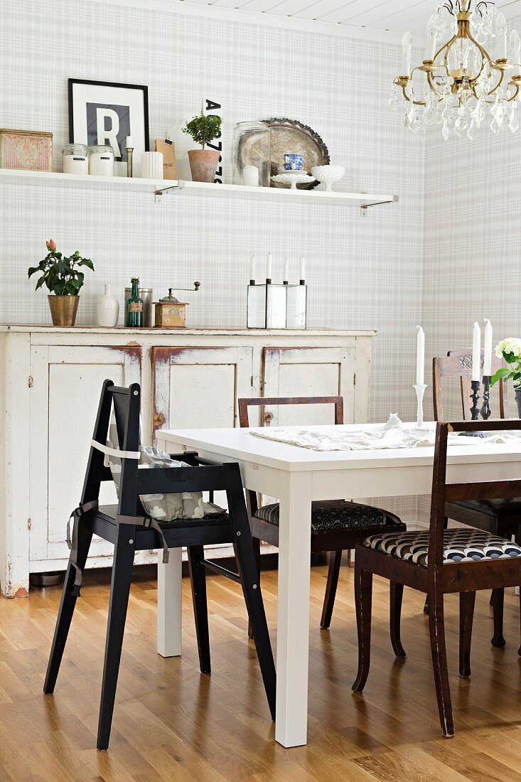 Black high chair at white table in front of vintage sideboard against grey ans white checked wallpaper