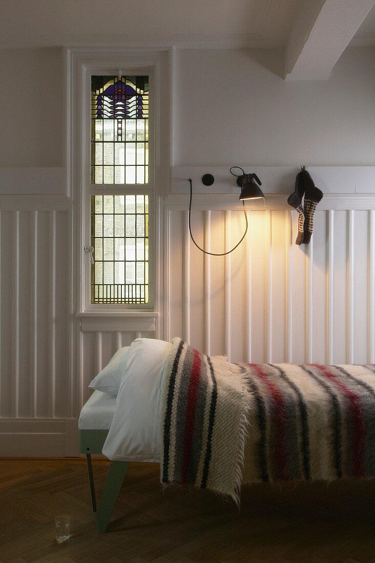 Single bed with striped bedspread, white-painted walls and wooden dado and window with Art Deco elements in traditional ambiance