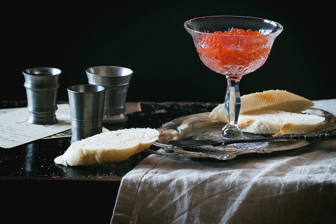 Roter Kaviar in Kristallschale, Weißbrot und Zinnbecher auf schwarzem Tisch