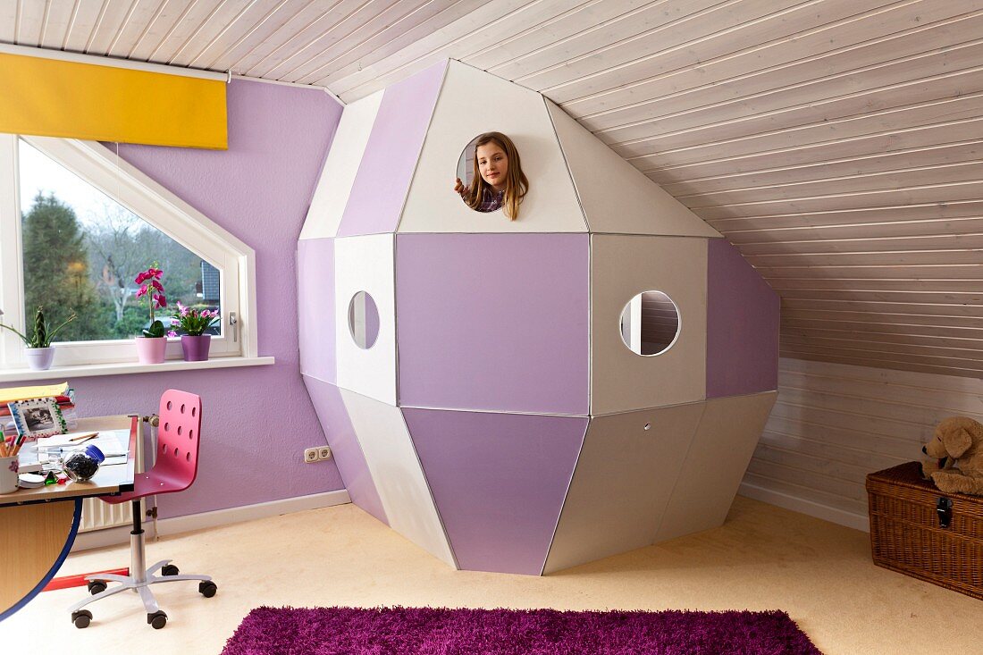 DIY space capsule made from white and purple wooden panels under sloping ceiling in child's bedroom; girl peeping through spy-hole