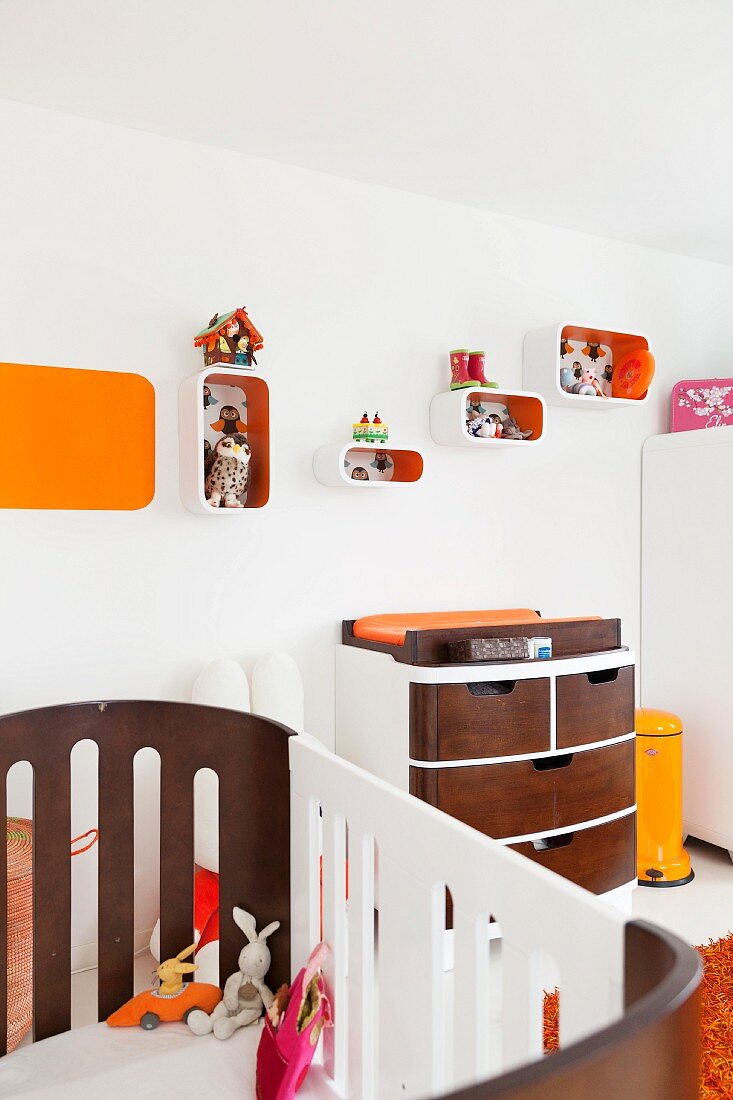 View across cot to custom chest of drawers with wooden fronts and shelving modules on wall with orange interiors