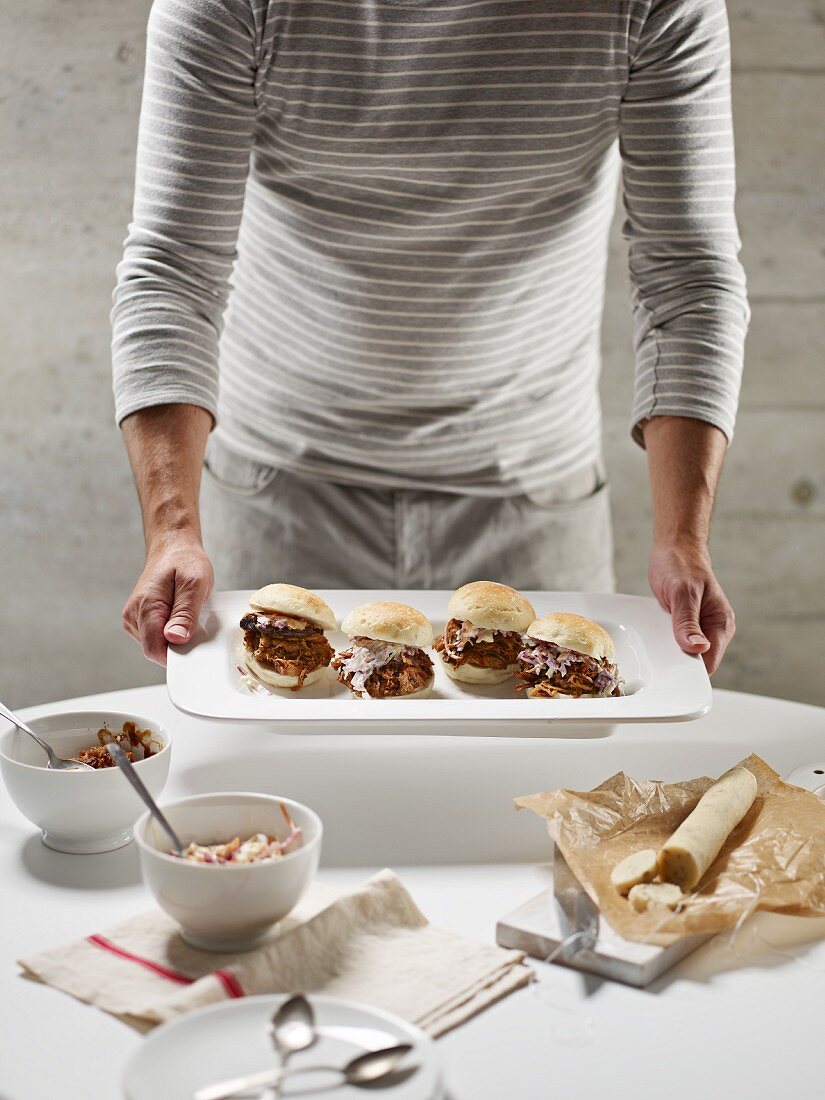 A man serving placing a platter of pulled pork burgers onto a table with pecan nut blue cheese shortbread pastry and bowls of salad