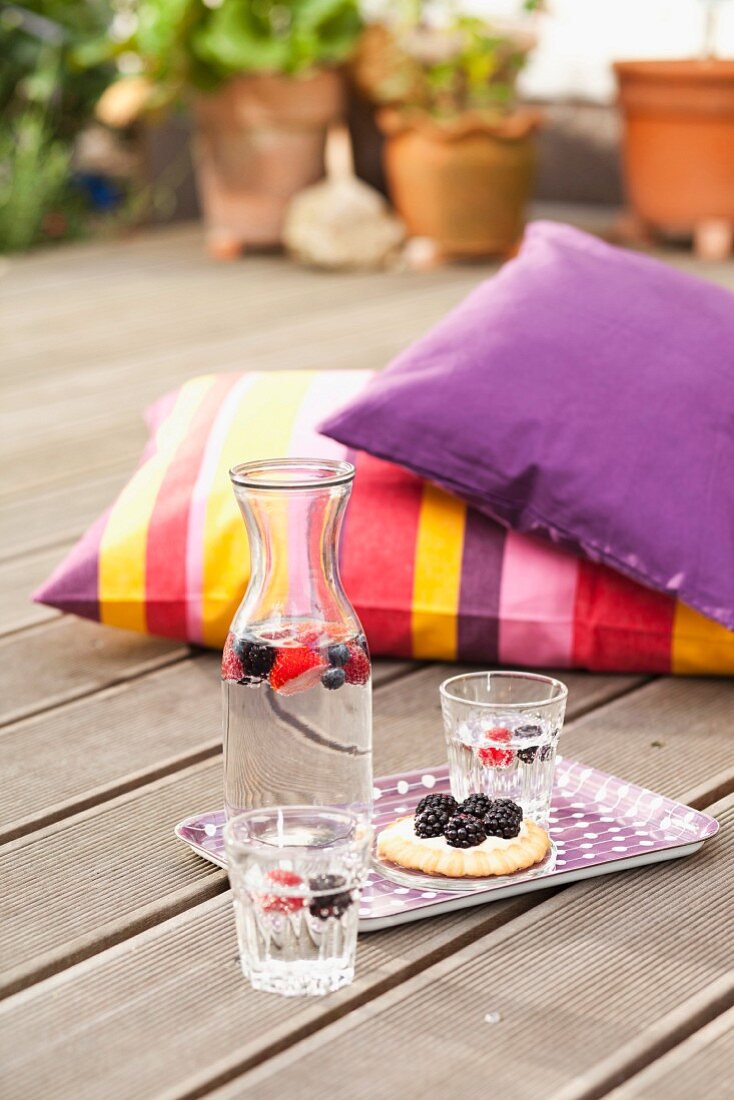 Tablett mit Erfrischungsgetränk, Gläsern und Törtchen auf Holzterrasse, im Hintergrund farbige Kissen