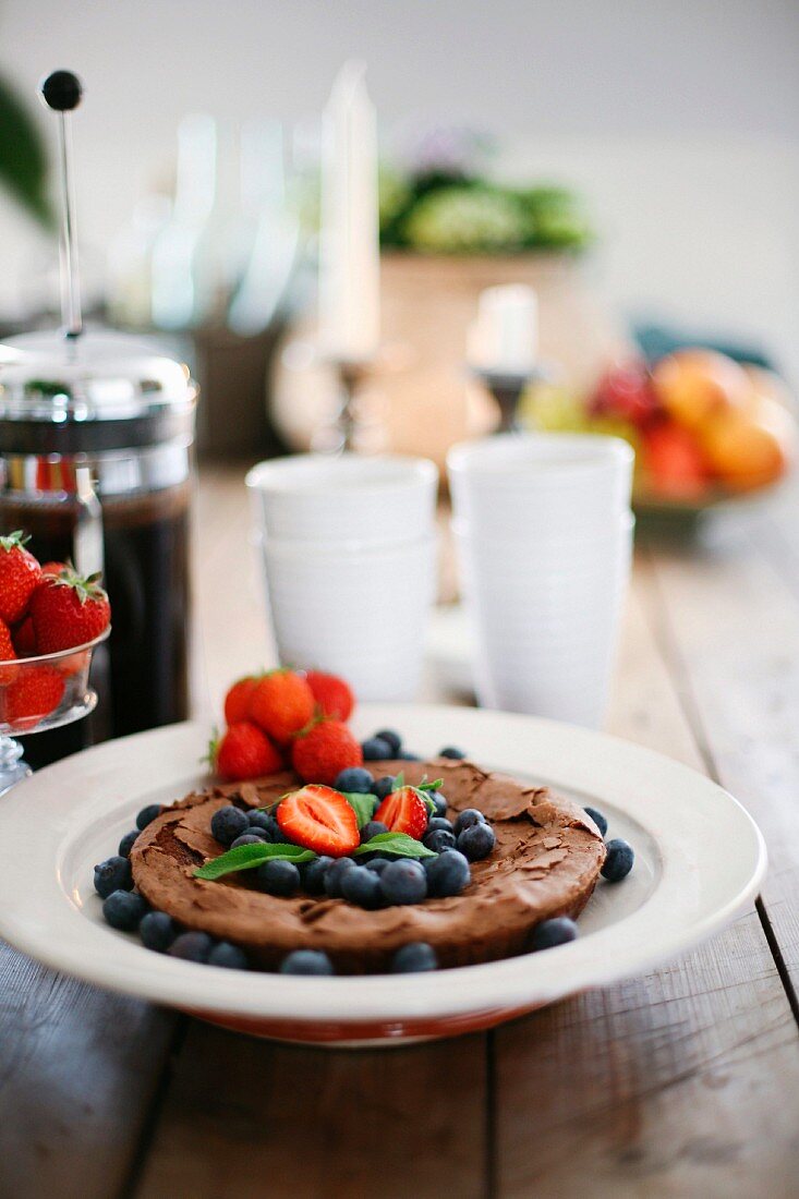 Chocolate cake with fresh blueberries and strawberries on a plate with white cups and a cafetière in the background