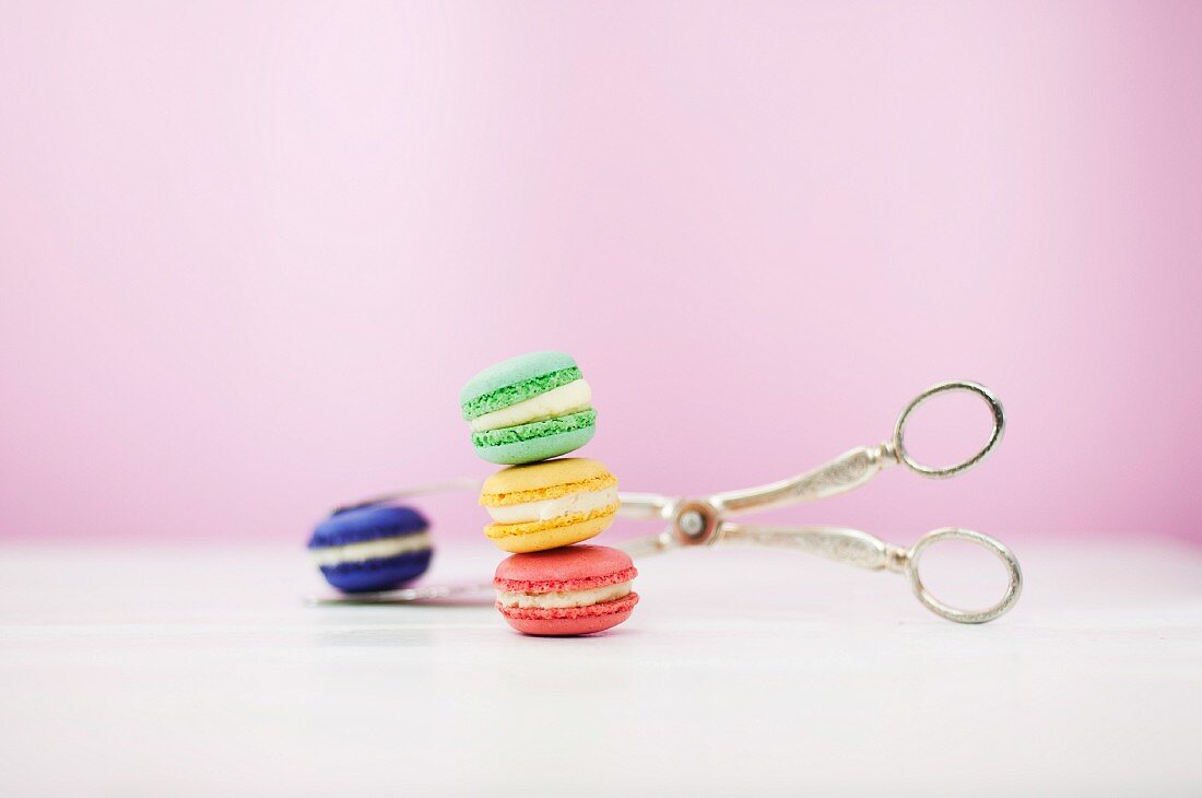 Colourful macaroons with a pair of silver tongs against a pink background