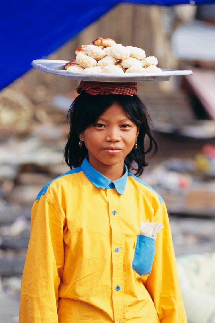 Mädchen mit gelbem Hemd eine Platte frisch gebackenes Gebäck auf dem Kopf tragend; Phnom Penh, Kambodscha