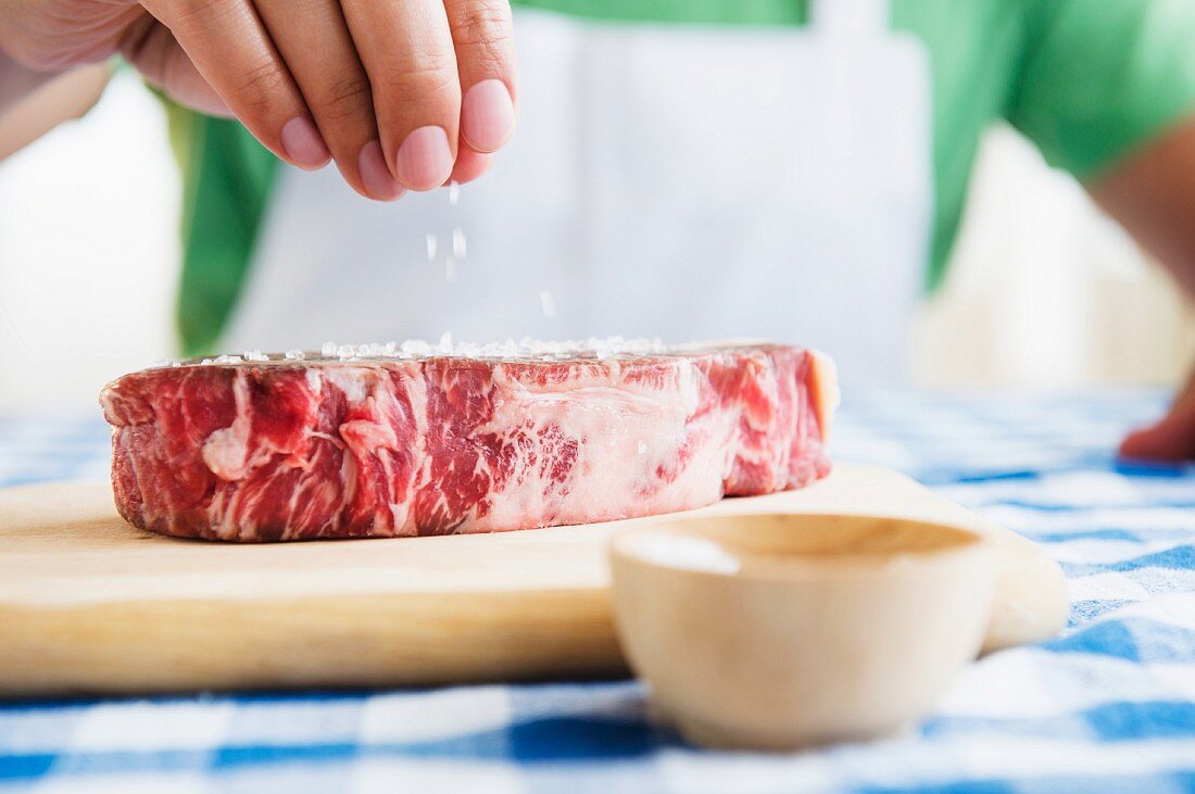 A raw beef steak being sprinkled with salt
